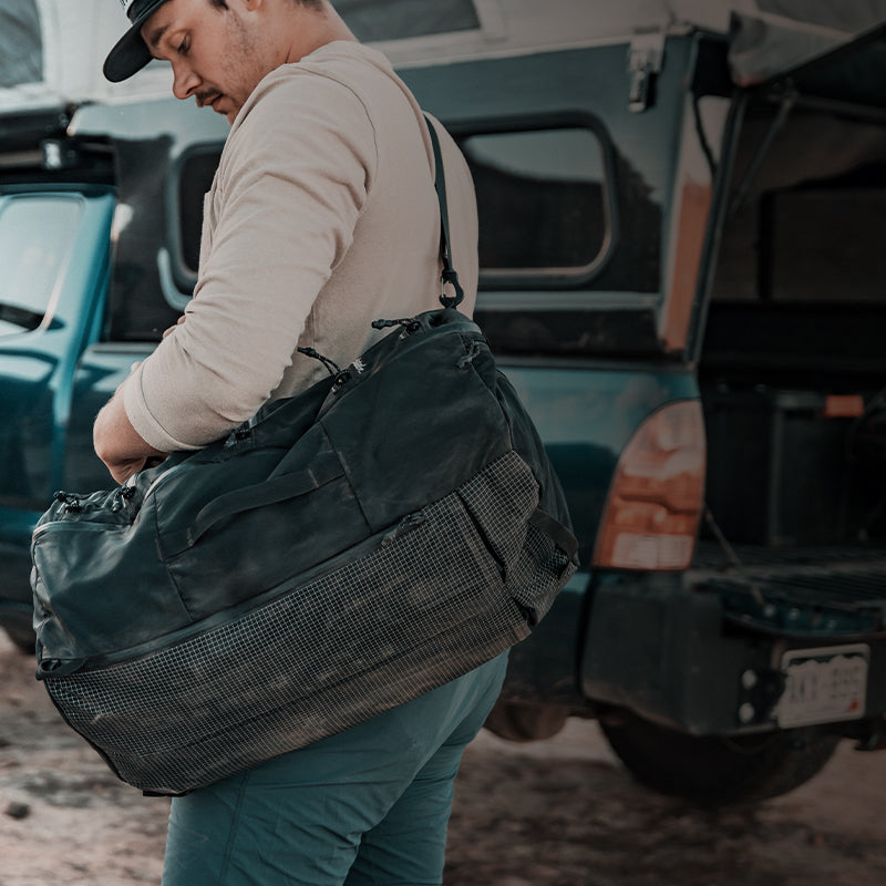 A person carrying a large black duffel bag stands near a vehicle, wearing a light shirt and cap.